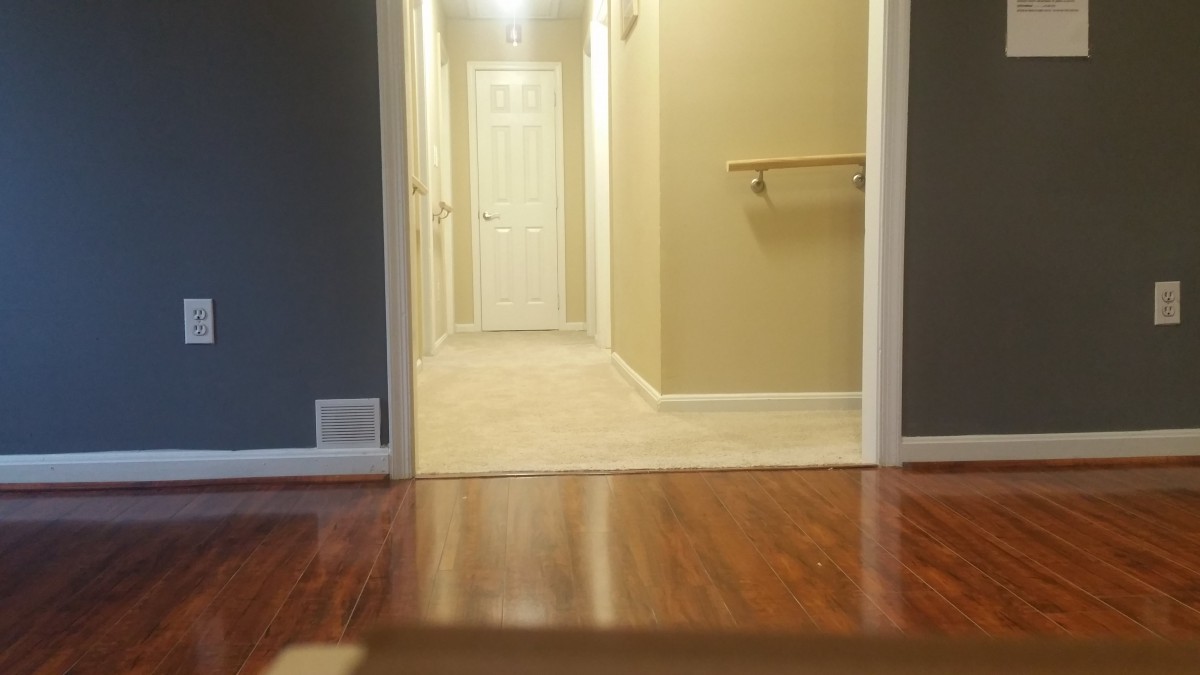 A hallway with a wooden floor leading to a closed door, featuring a handrail and illuminated by ceiling lights.