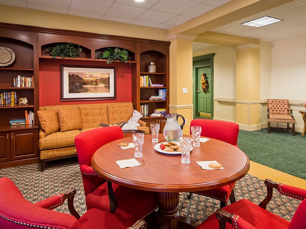 A cozy meeting room with a round table, red chairs, and a comfortable couch, featuring artwork and a bookshelf.