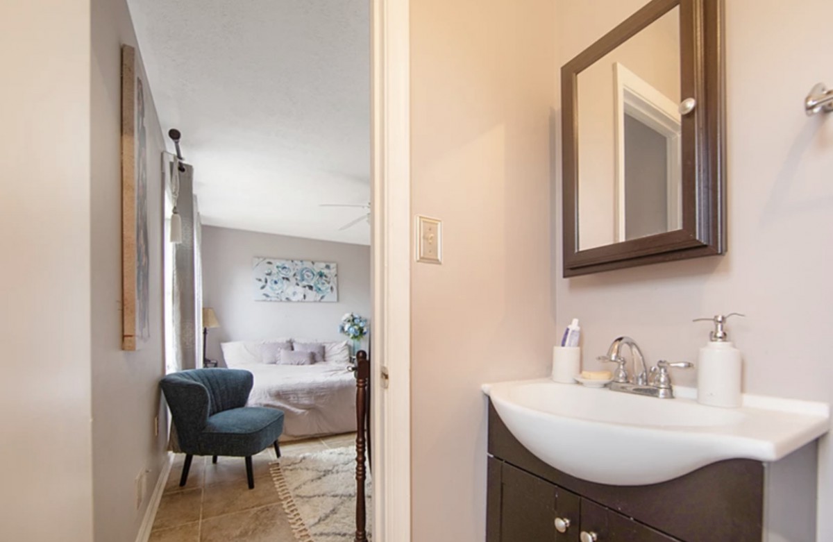 A view from a bathroom with a sink and mirror into an adjacent bedroom featuring a chair and bed.