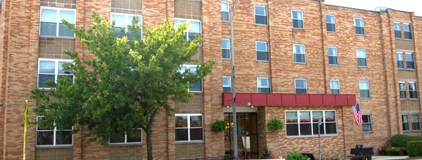 A brick apartment building with multiple windows and a small entrance canopy, surrounded by greenery.