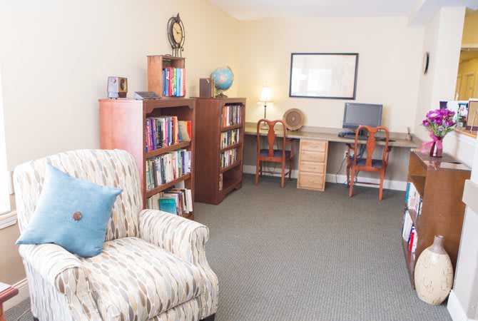 A cozy living room with bookshelves, an armchair, and a desk area featuring a computer and lamp.