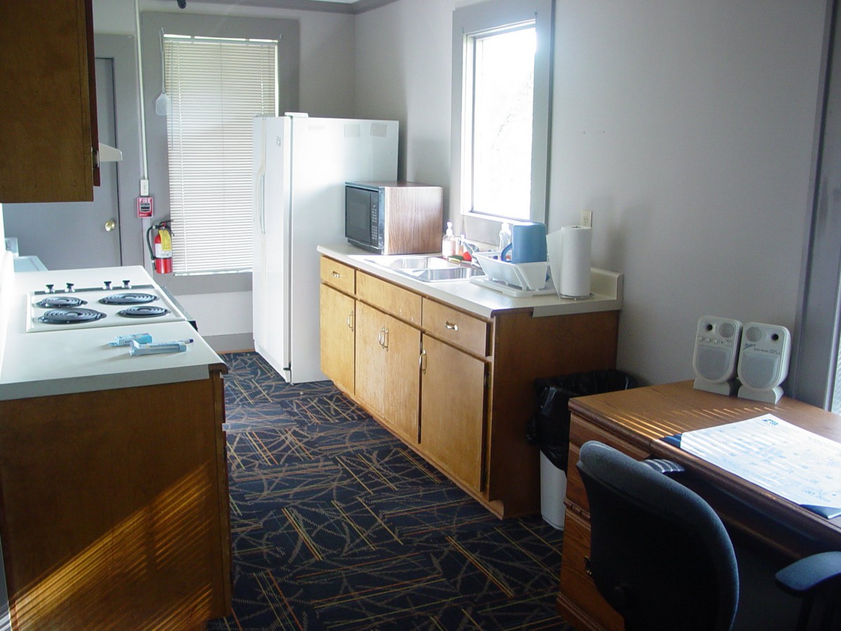 A modest kitchen area with wooden cabinets, a refrigerator, and a small desk with a chair near the window.