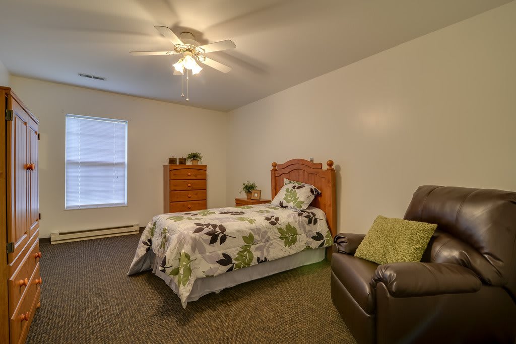 A cozy bedroom with a single bed, wooden dresser, and armchair, featuring a ceiling fan and carpeted floor.