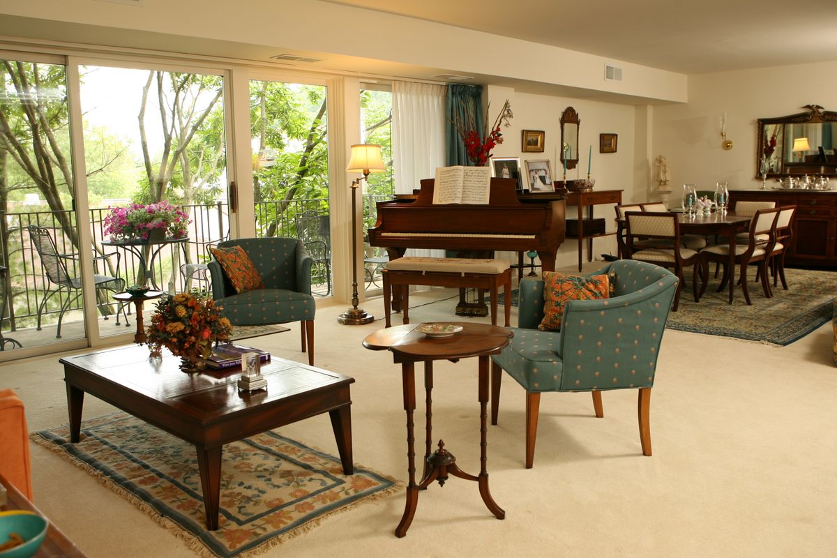 A cozy living room with elegant furniture and a grand piano, featuring natural light from large windows.
