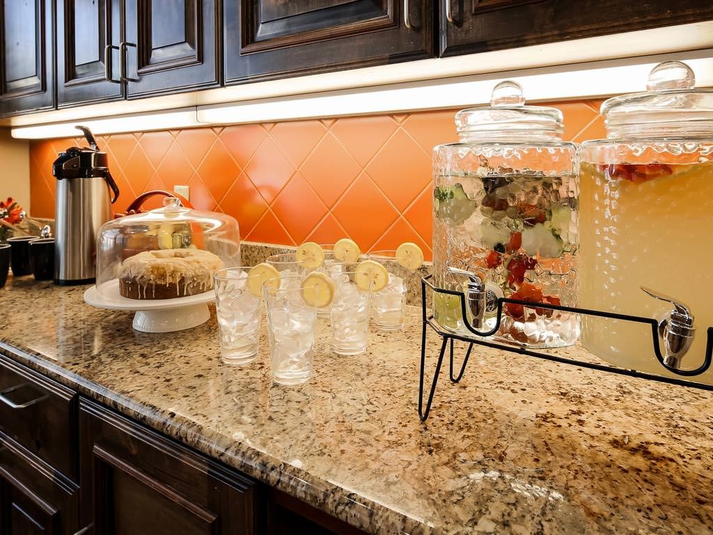 A kitchen counter featuring a beverage dispenser with infused water, glasses garnished with lemon slices, and a cake under a glass dome.