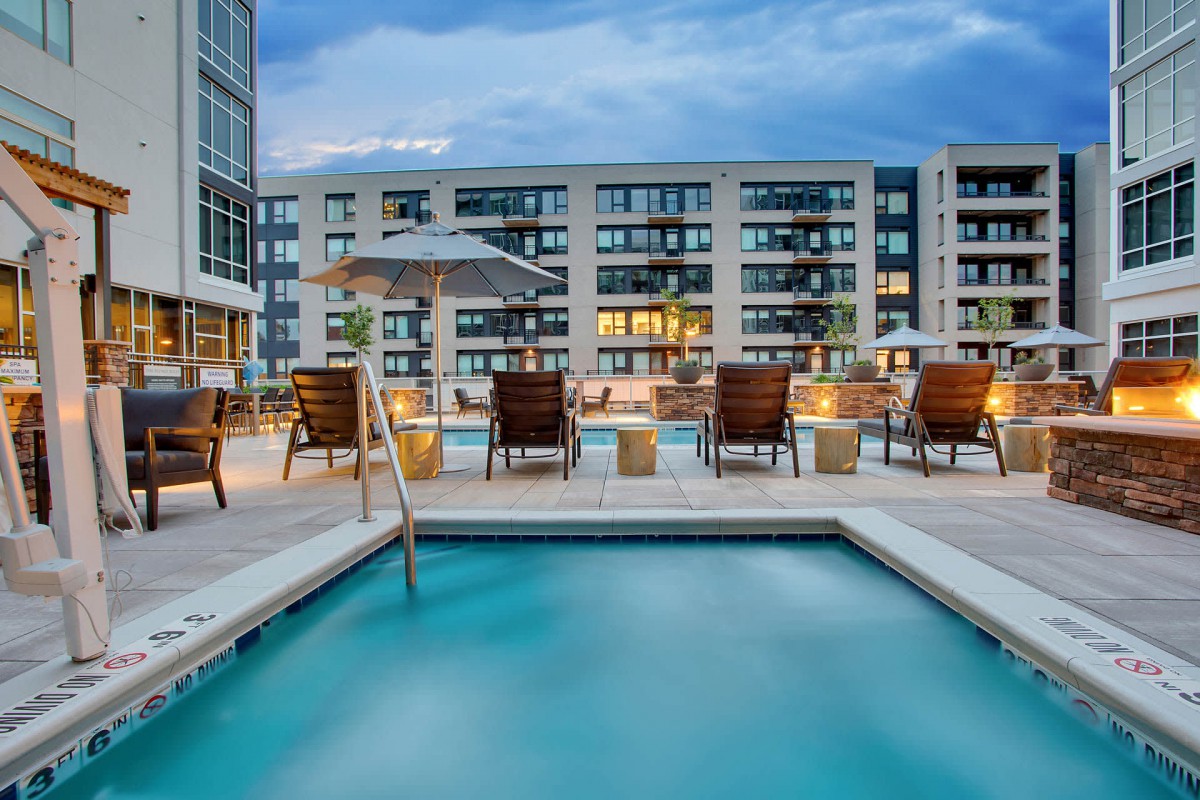 A rooftop swimming pool area with patio chairs and umbrellas in an apartment complex during the evening.