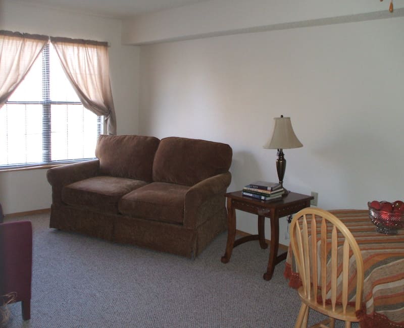 A cozy living room with a brown sofa, side table, and lamp near a window with light curtains.