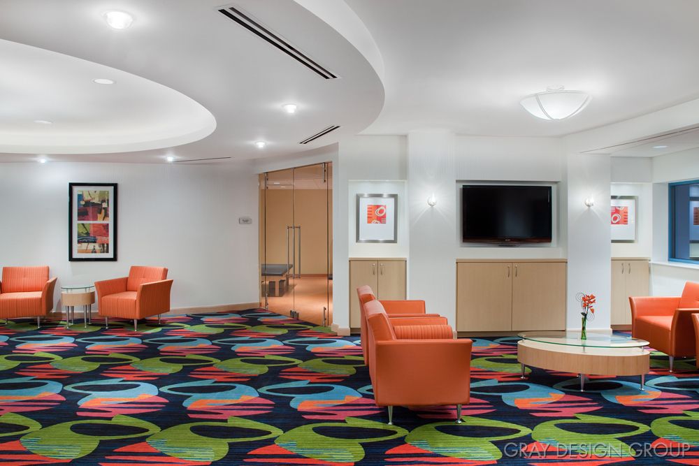 A modern lobby with colorful carpeting, orange chairs, a television, and artwork on the wall.