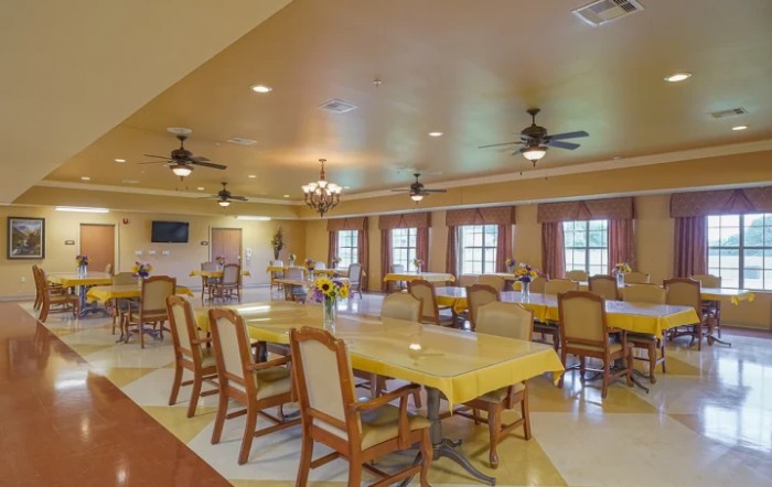 A spacious dining area with multiple tables and chairs, illuminated by ceiling lights and natural light from large windows.
