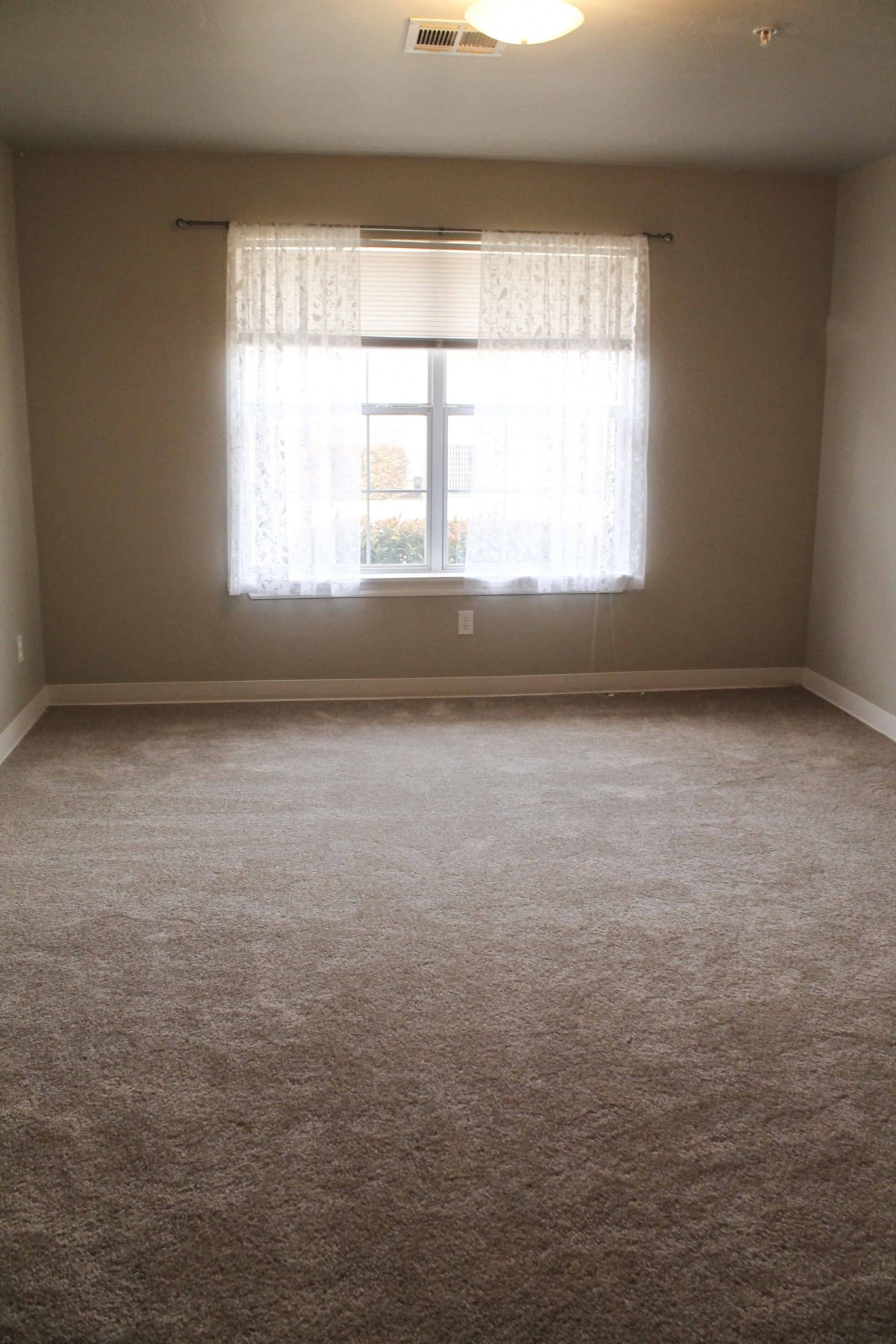 An empty room with beige carpeting and a window covered by sheer curtains.