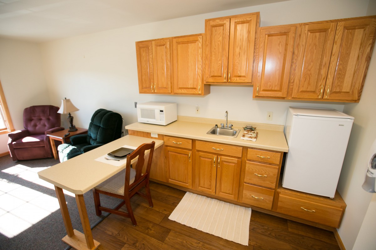 A compact kitchen area featuring wooden cabinets, a countertop with a microwave, sink, and fridge, alongside a small table and chair.