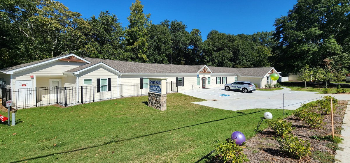 A single-story building with a driveway and a car parked in front, surrounded by grass and trees under a clear blue sky.