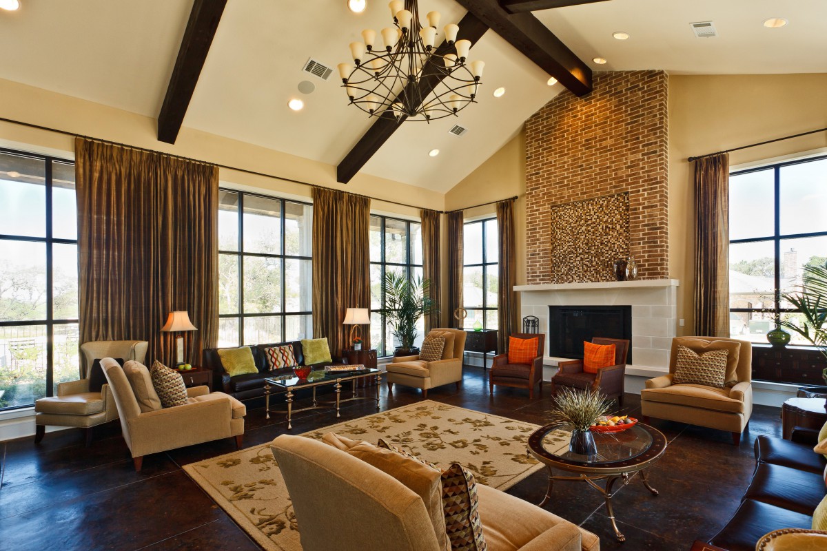 A spacious living room featuring traditional decor, high ceilings with exposed beams, and a large chandelier. Comfortable seating surrounds the fireplace, complemented by large windows that bring in ample natural light.