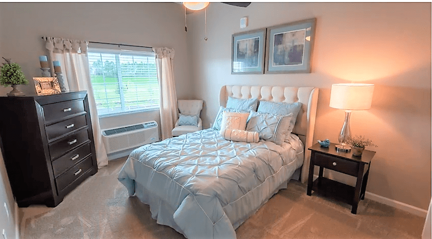 A cozy bedroom with a neatly made bed, featuring soft pillows and blankets, flanked by a small chair and a nightstand with a lamp. Natural light streams in through the window, creating a warm and inviting atmosphere.