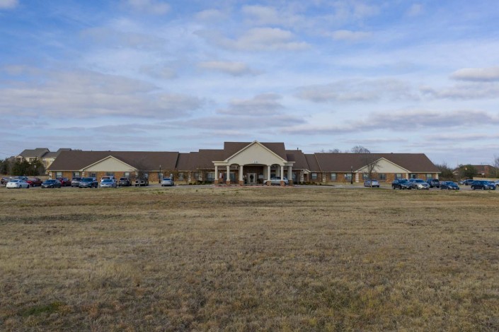 A large building with a field in the foreground, flanked by parked cars under a partly cloudy sky.
