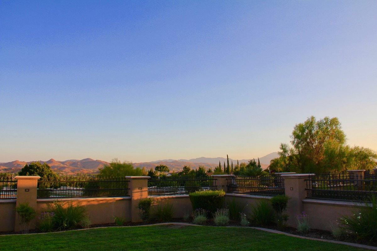 A serene landscape with a view of mountains under a clear evening sky, framed by trees and a stone fence.