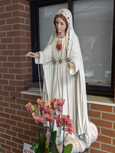 A serene statue of Mary adorned with a rosary, surrounded by vibrant flowers against a brick backdrop.