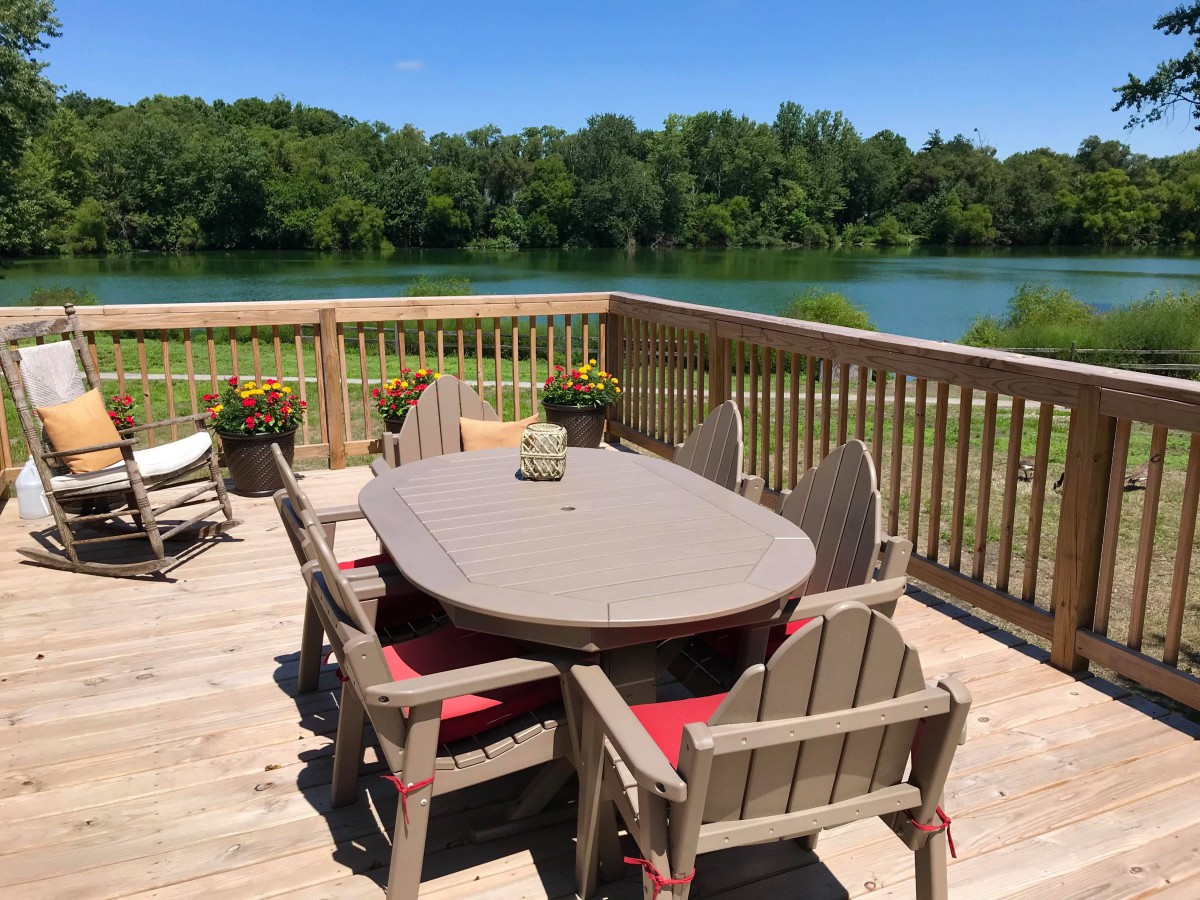 A wooden deck with outdoor furniture overlooks a serene lake surrounded by lush greenery.