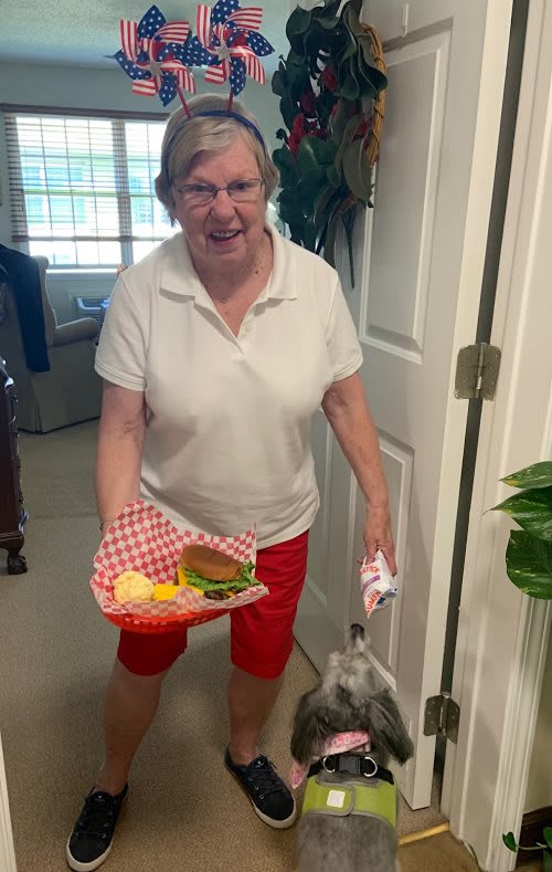 A woman in red shorts and a white shirt is smiling while holding a tray with a burger and chips indoors.