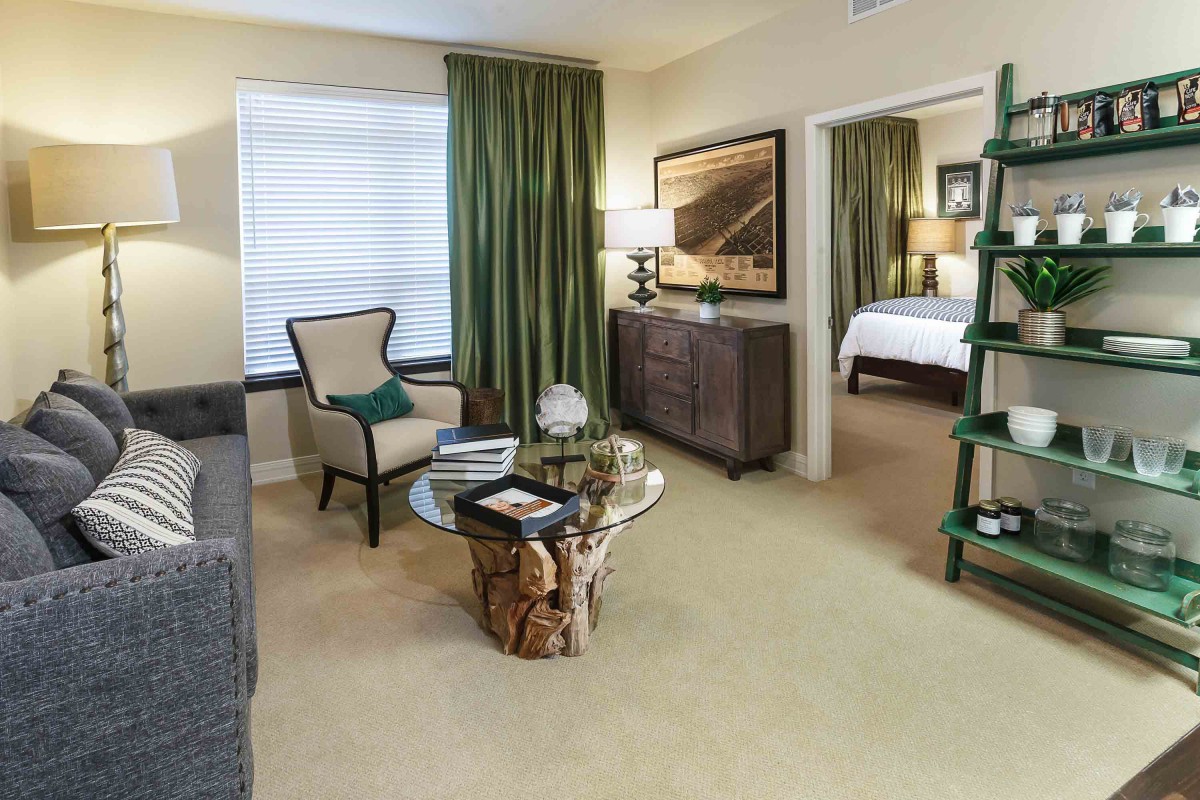A cozy living room with green curtains, stylish furniture, and a view into the adjacent bedroom.