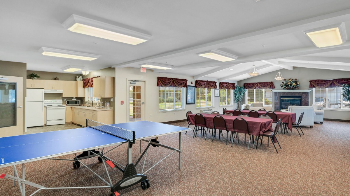 A bright community room featuring a ping pong table, dining setup, and large windows.