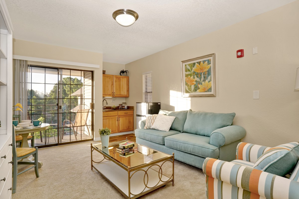 A cozy living room with a blue sofa, a glass coffee table, and a small kitchenette.