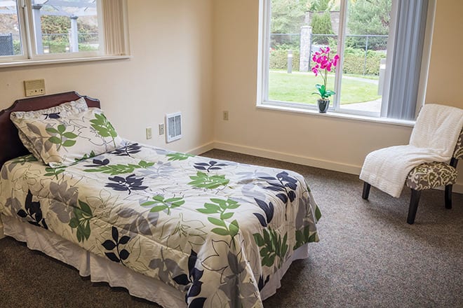 A simple and cozy bedroom with a single bed, floral bedding, a window view, and a flower vase on the sill.