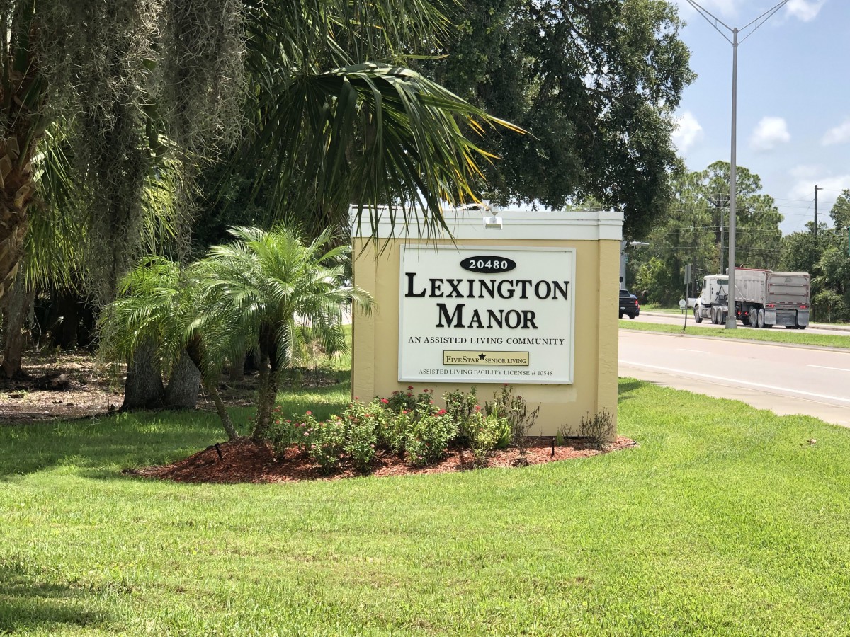 A roadside sign for Lexington Manor surrounded by palm trees and well-maintained greenery.