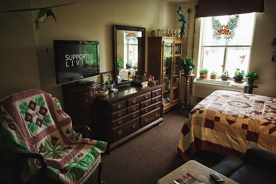 A cozy bedroom with a dresser, TV, and chair, featuring a quilt-covered bed and several plants by the window.
