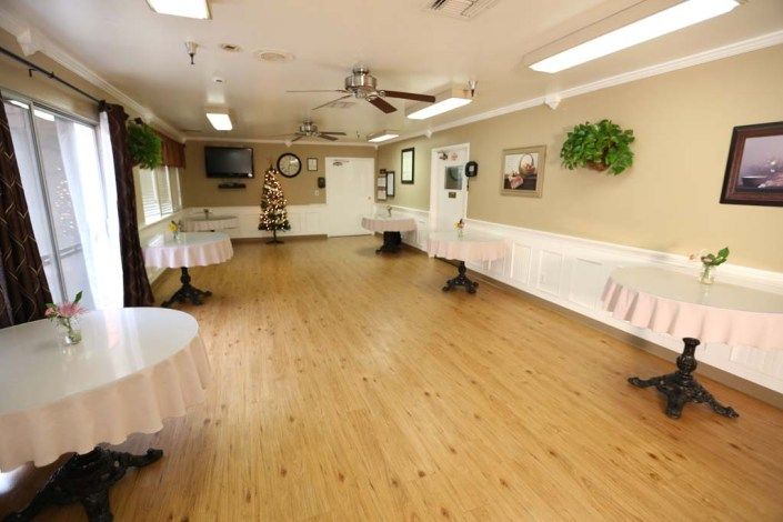 An empty indoor hall with wooden flooring, round tables with white tablecloths, decorative plants, and ceiling fans.