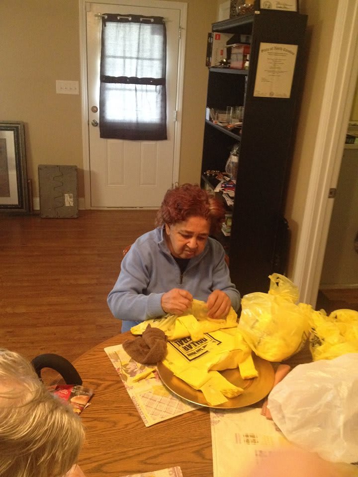 A woman is seated at a table in a kitchen, working on a crafting project with yellow materials.