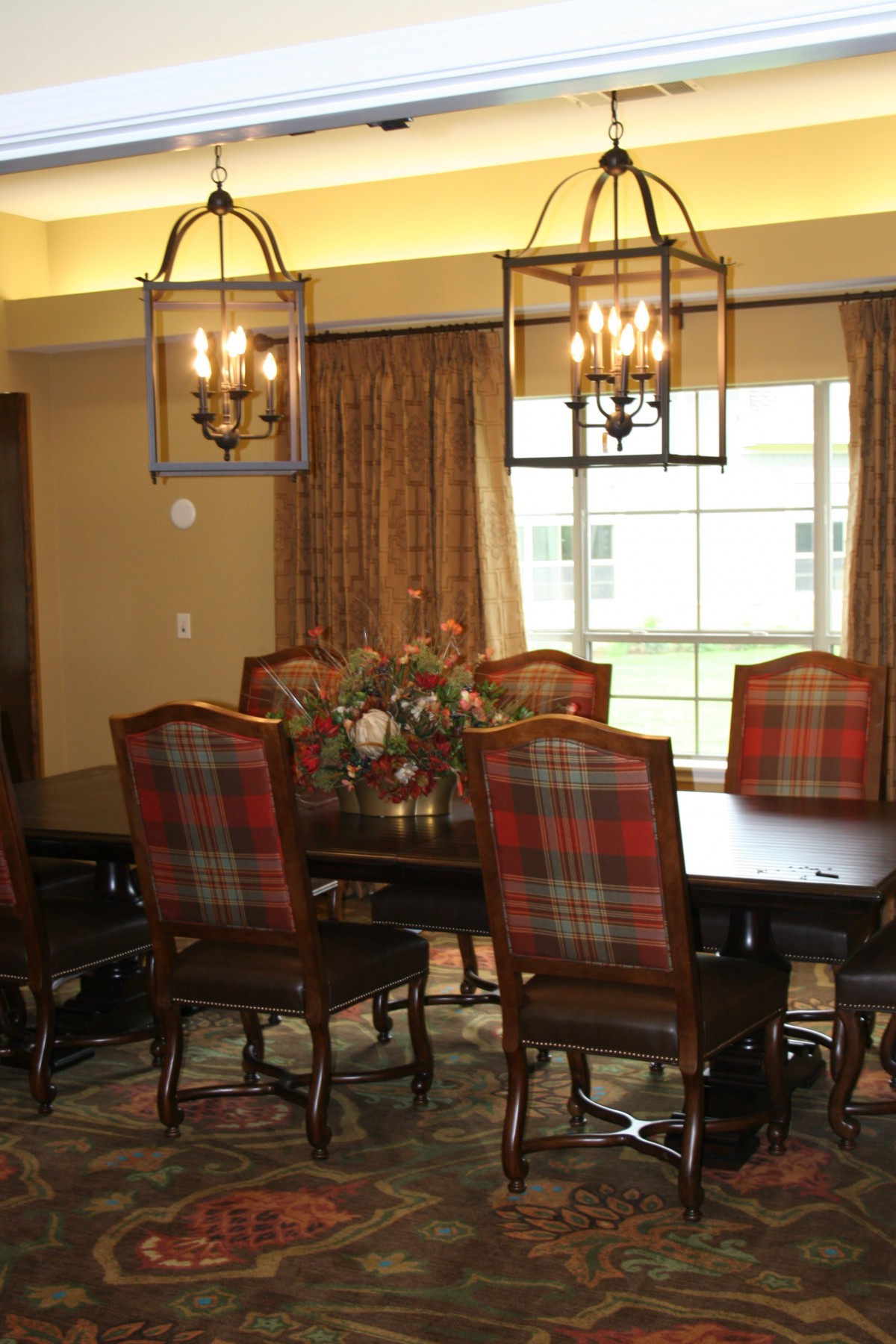 A cozy dining room featuring a large wooden table with plaid upholstered chairs, two elegant light fixtures, and a window adorned with curtains.