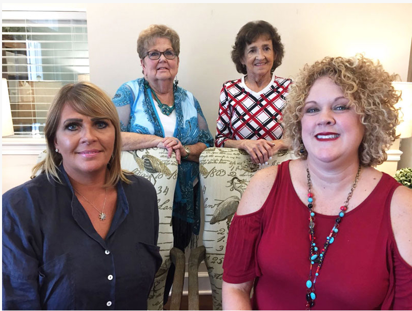 A group of four women smiling indoors, dressed in casual attire.