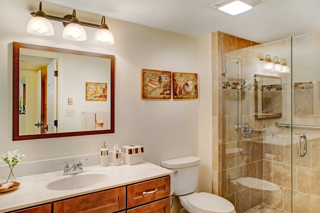 A well-lit bathroom featuring a shower, toilet, and sink with wooden cabinetry and decorative wall art.