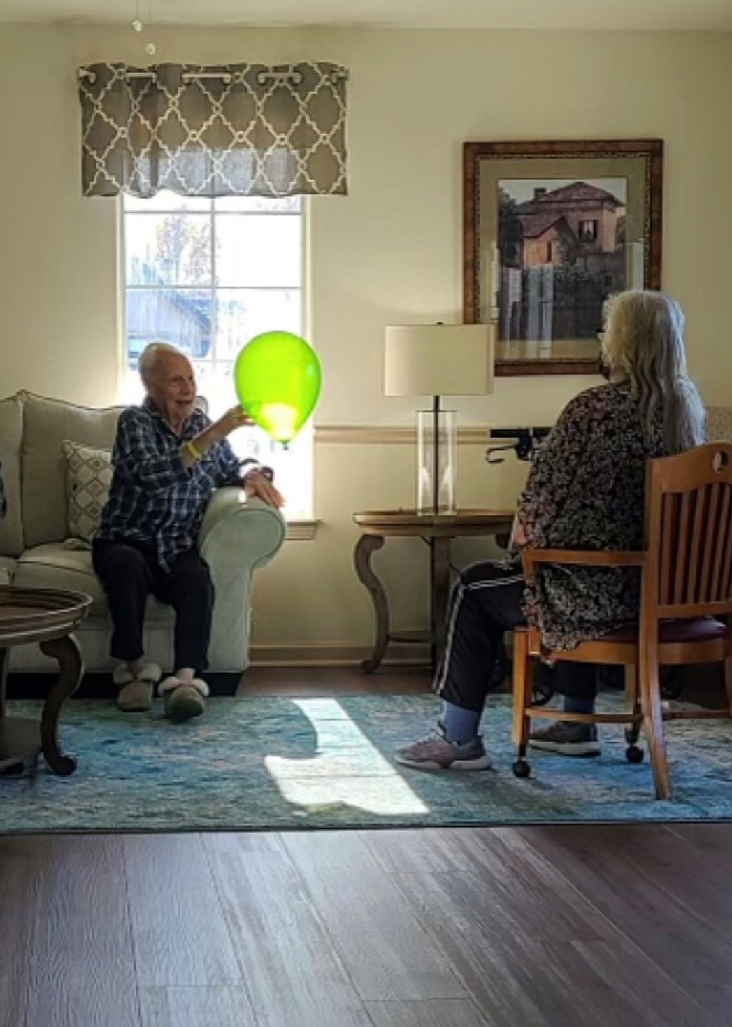 Two seniors are seated in a living room, engaging with a green balloon.