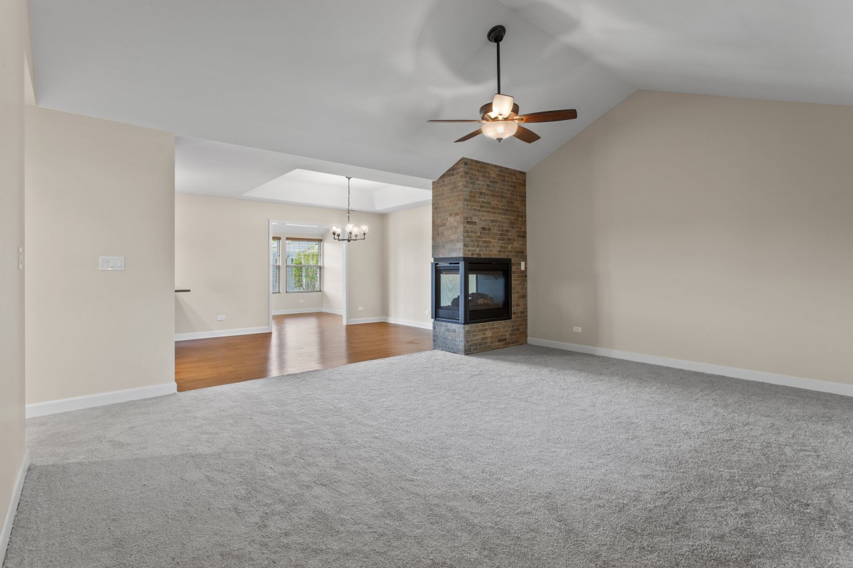 A spacious living room with a central fireplace and ceiling fan, featuring carpeted flooring and ample natural light.