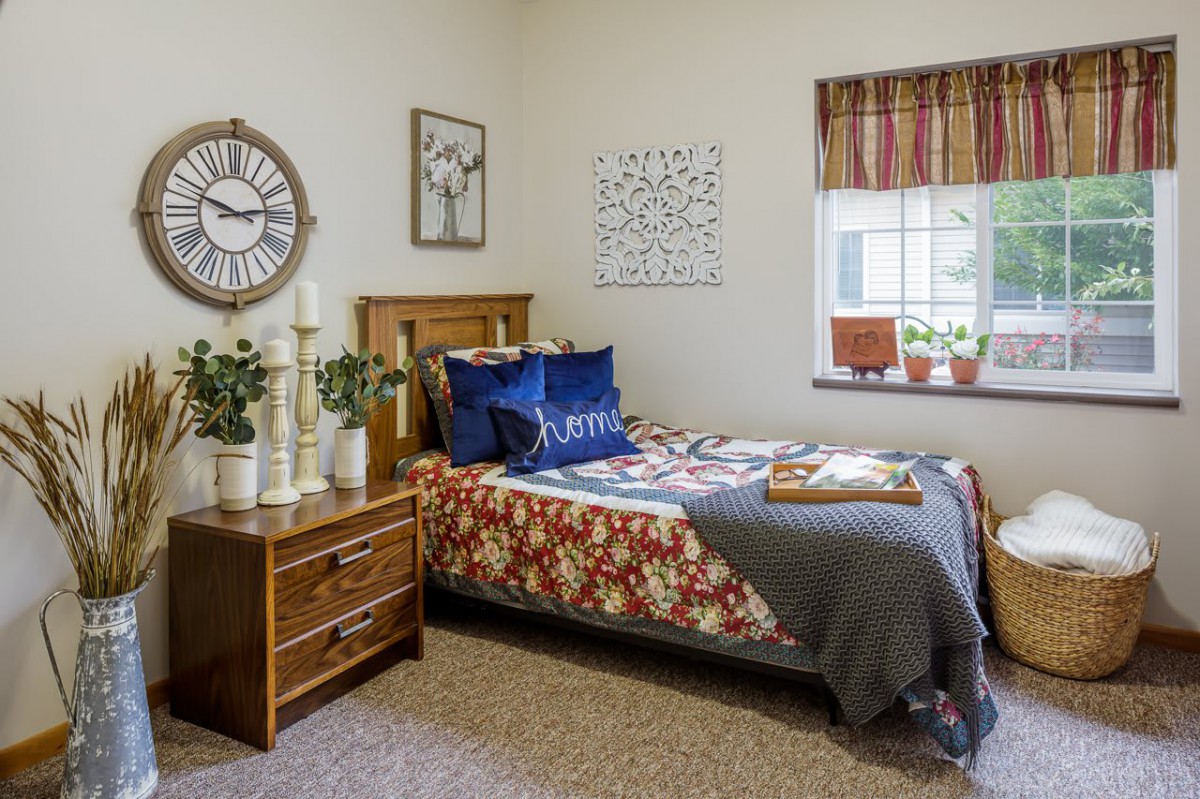 A cozy bedroom with a wooden bed, colorful bedding, and decorative plants. A large wall clock and artwork adorn the walls.