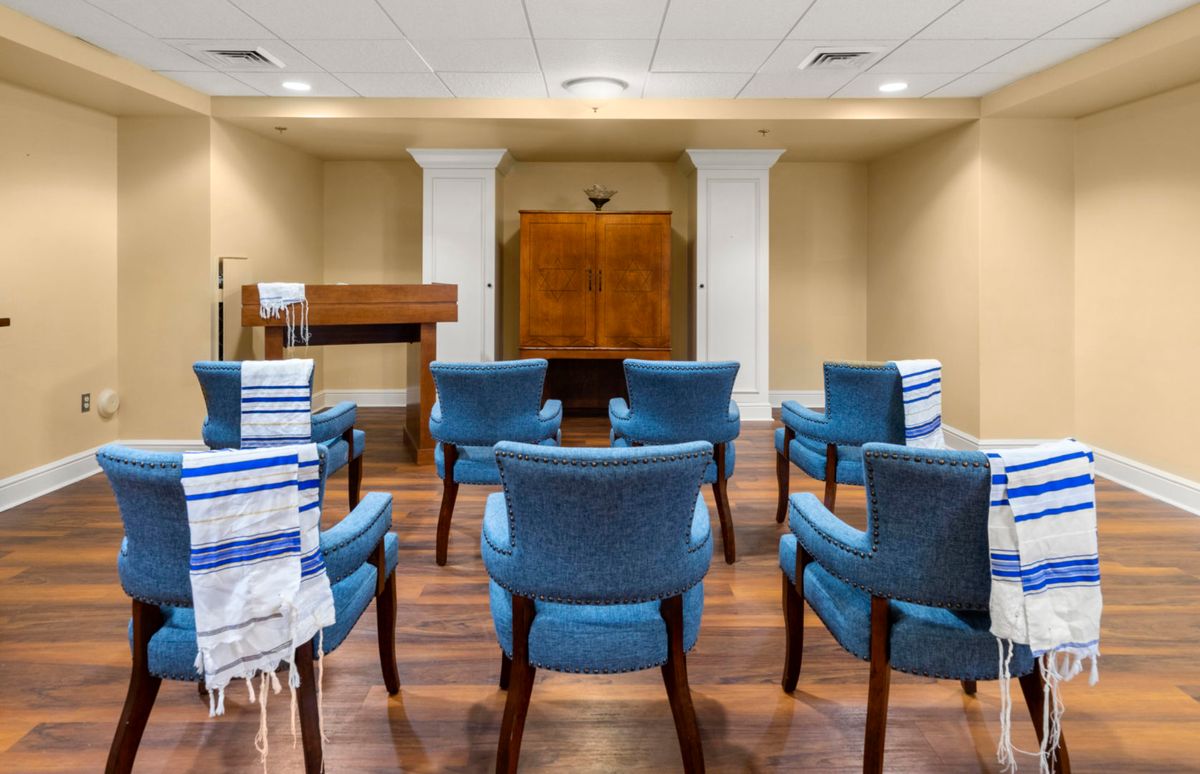 A small synagogue room with chairs arranged in rows, a podium, and prayer shawls draped over some of the chairs.