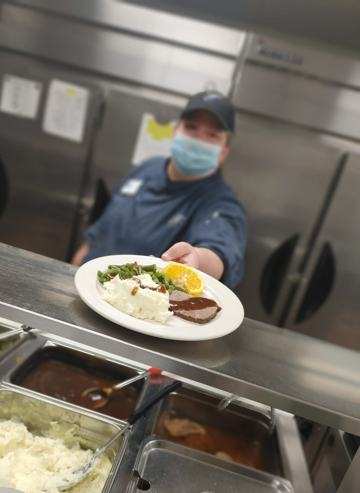 A chef wearing a mask presents a plated meal in a restaurant kitchen.