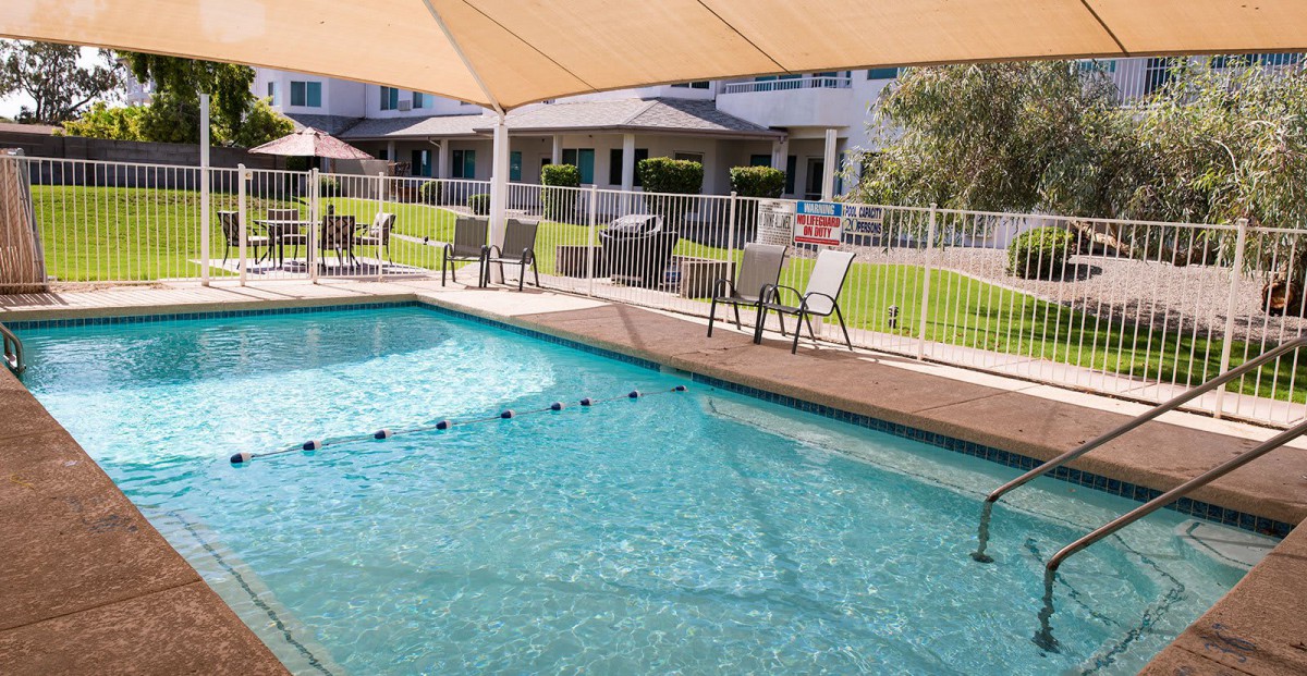 An outdoor swimming pool area with shaded seating and a surrounding fence.