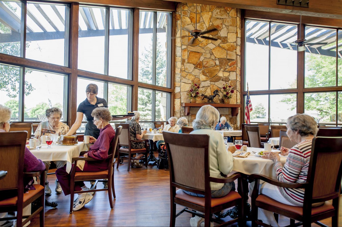 Seniors dining in a well-lit restaurant with large windows and a stone fireplace.