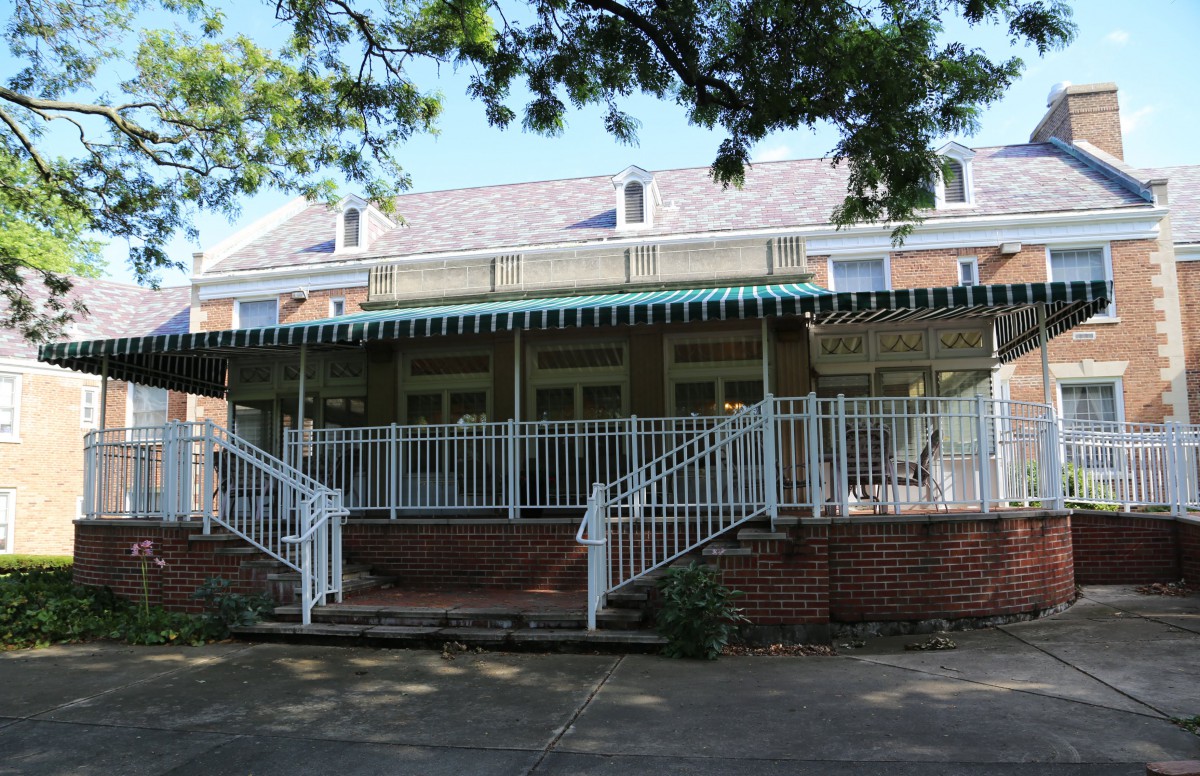 A residential house with a large porch, featuring brick steps and white railings, shaded by trees.