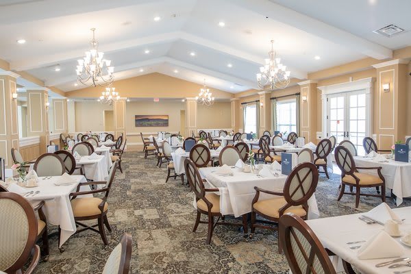 An elegant dining room with neatly arranged tables and chairs, featuring chandeliers and large windows.