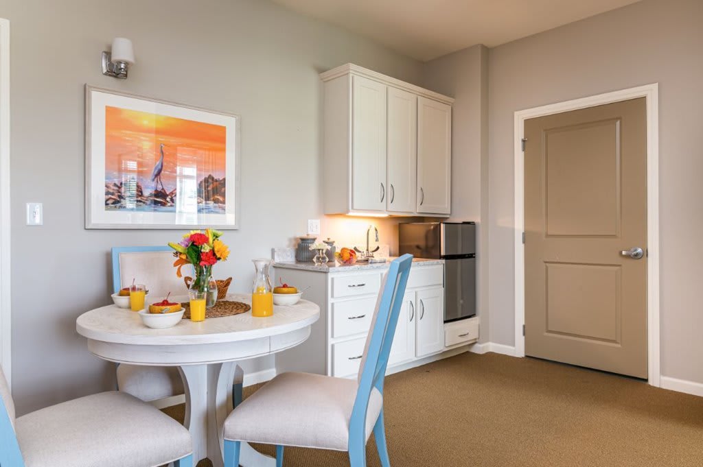A cozy kitchen and dining area featuring white cabinets, a small round table with chairs, and a colorful fruit bowl.