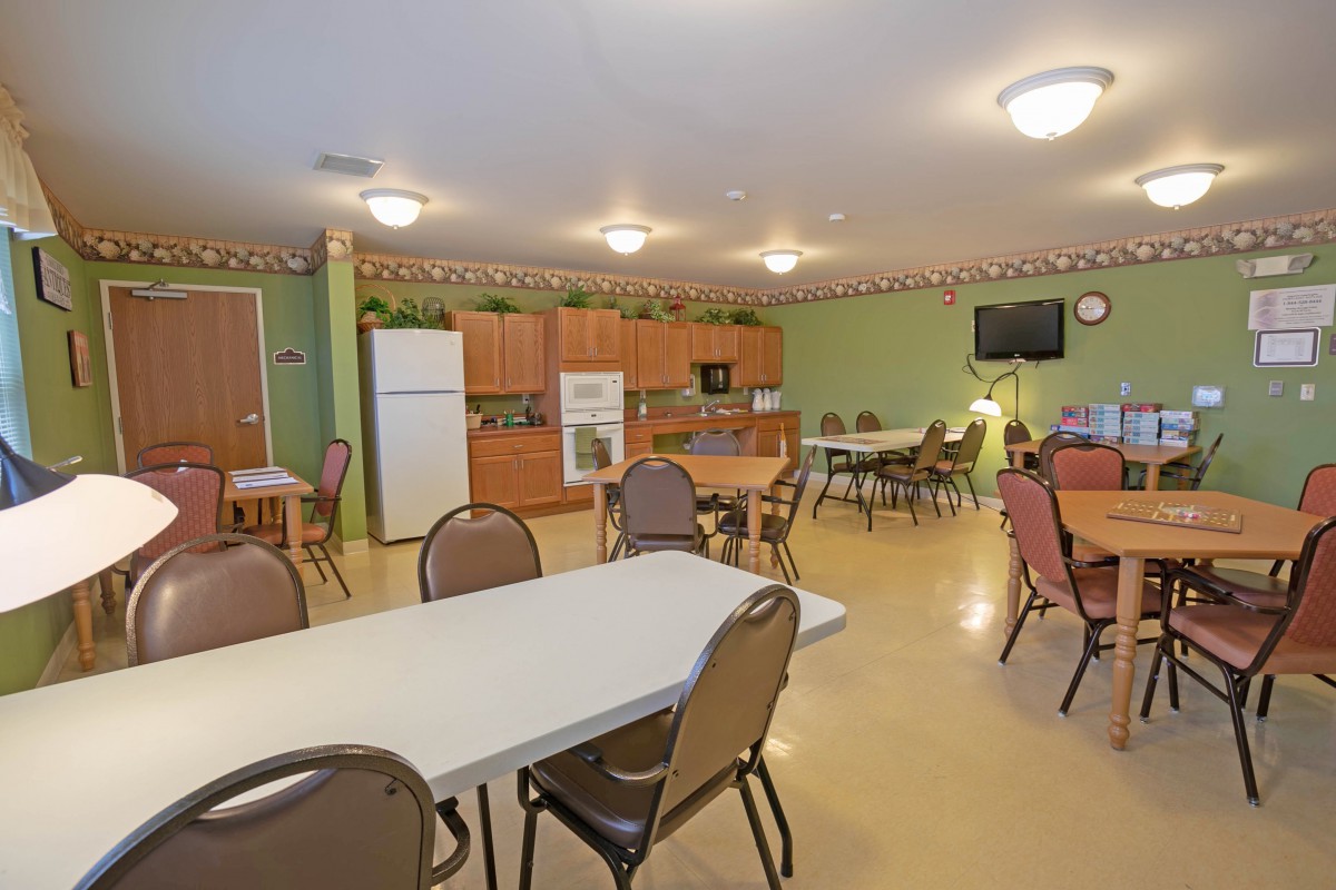 A community room featuring a kitchen area with tables and chairs, equipped with a refrigerator, microwave, and television.