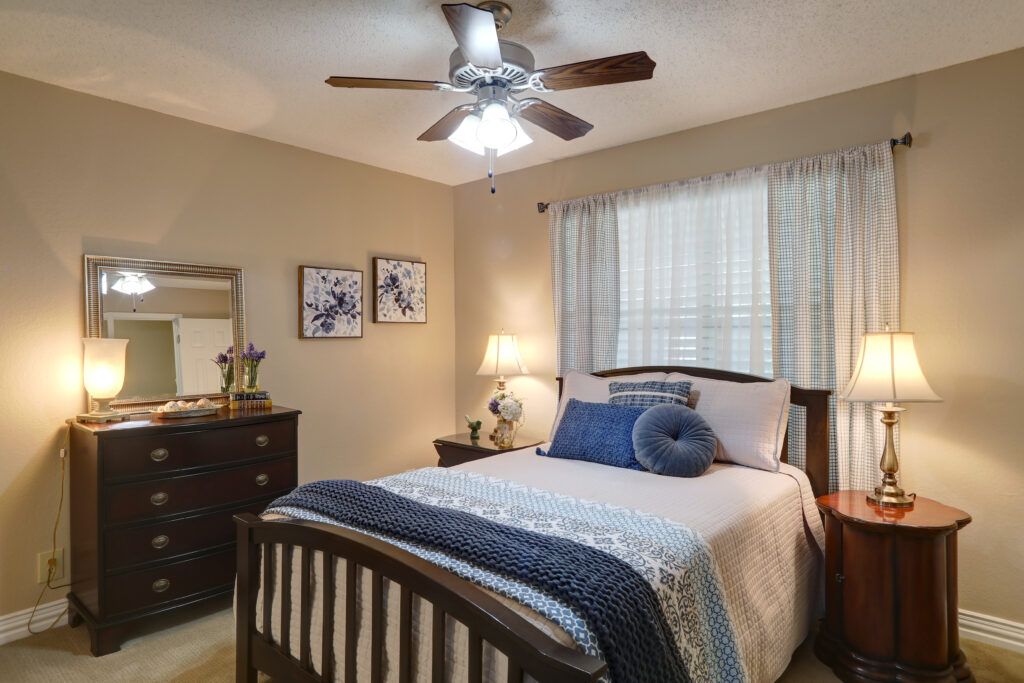 A cozy bedroom with a neatly made bed, featuring blue accents, a ceiling fan, and warm lighting.