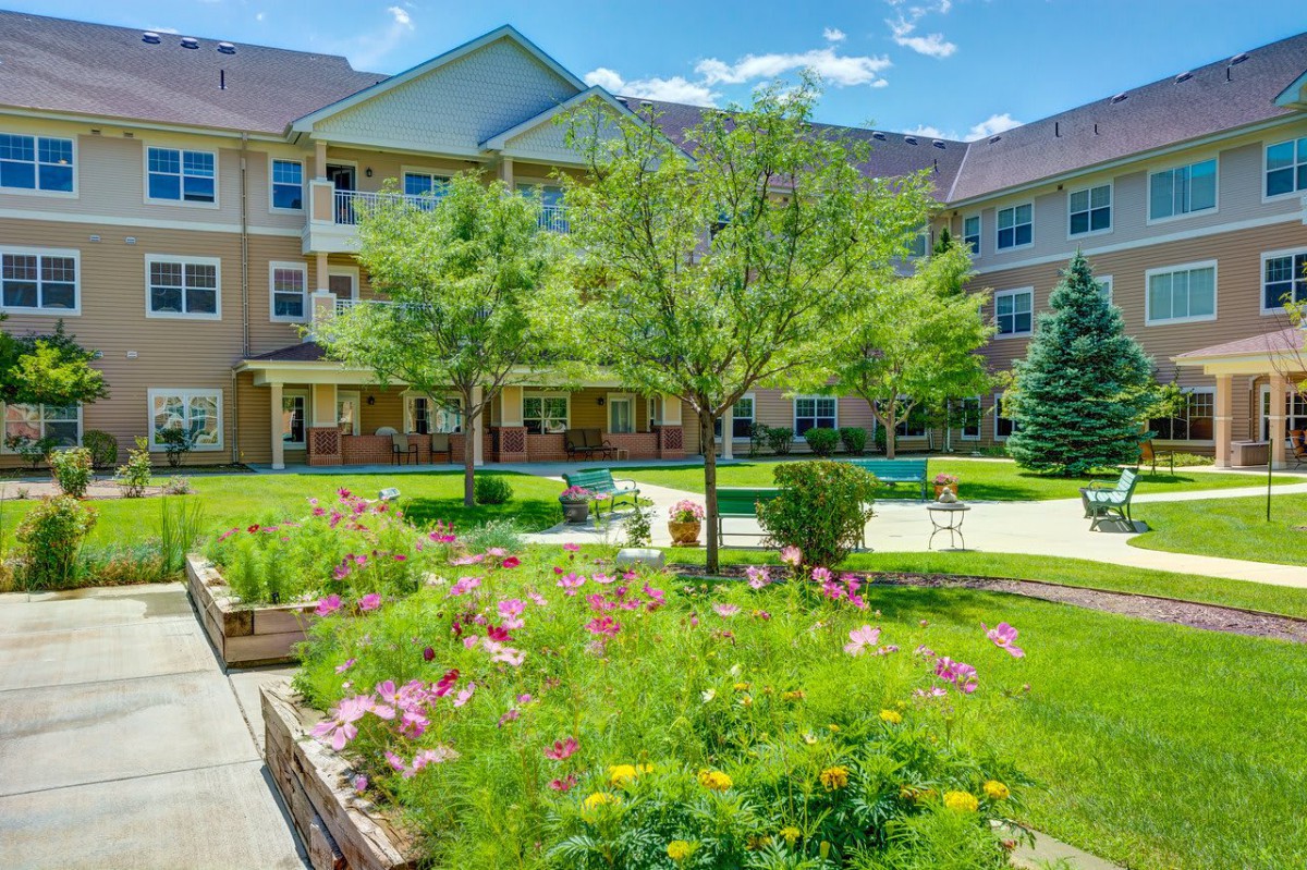 A beautiful courtyard garden with lush greenery and colorful flowers in front of a multi-story building under a bright blue sky.