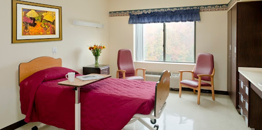 A hospital patient room featuring a bed, chairs, and a window with floral arrangements.