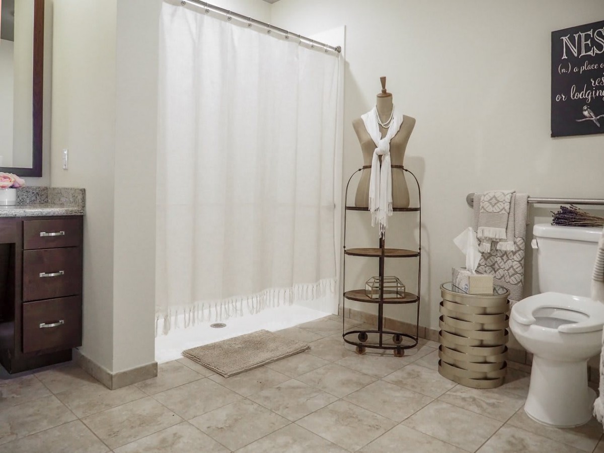 A clean and modern bathroom featuring a white shower curtain, neatly arranged towels on shelves, and a mannequin used for decoration.