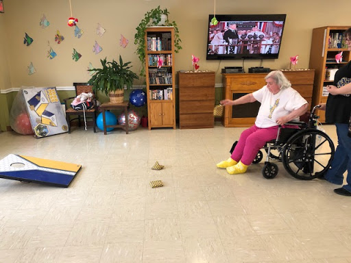 An elderly woman in a wheelchair participates in a game activity in a cheerful indoor setting.